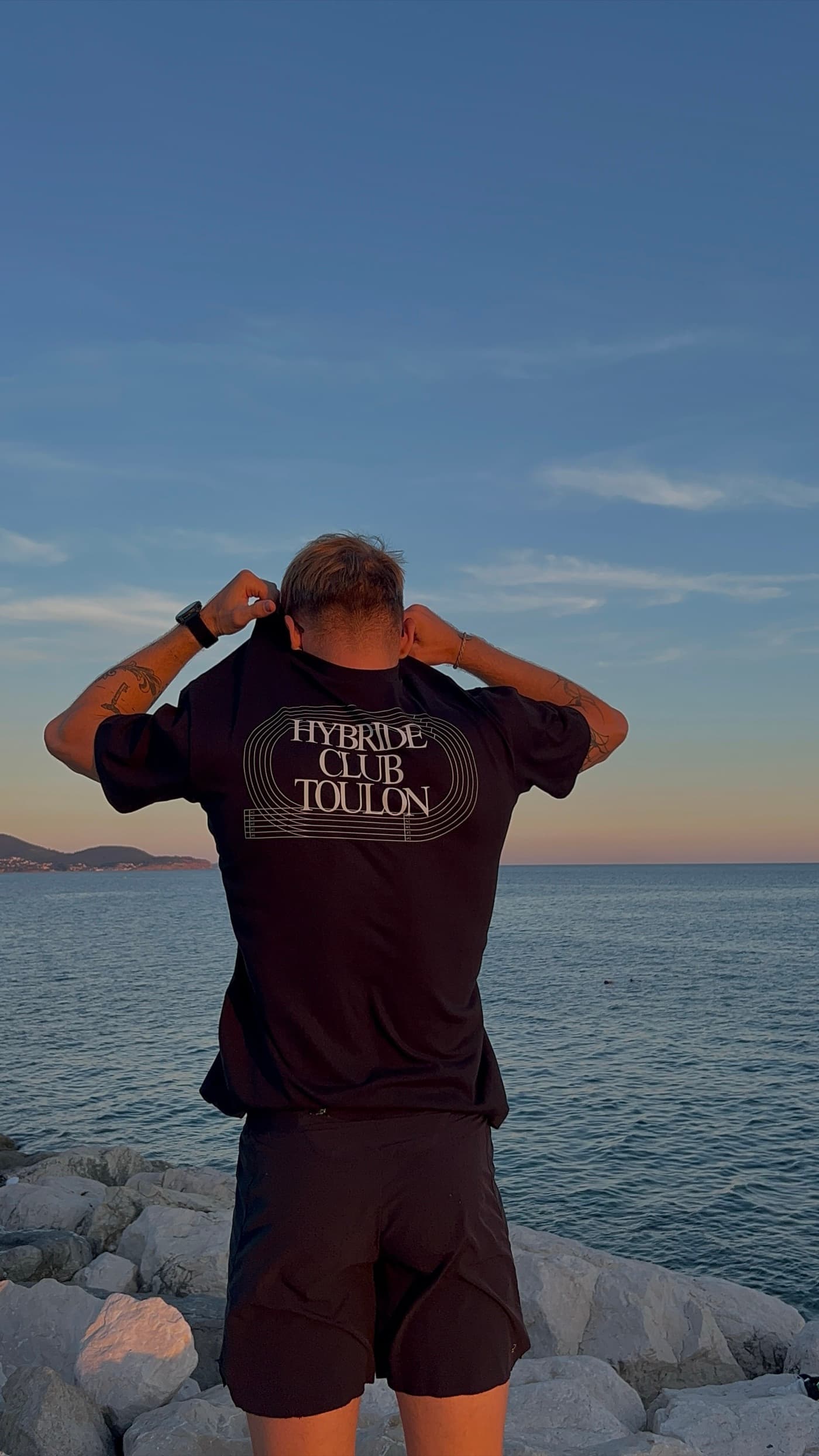 Un membre du club de dos sur les rochers, ajustant sa casquette, tee-shirt noir floqué Hybride Club Toulon
