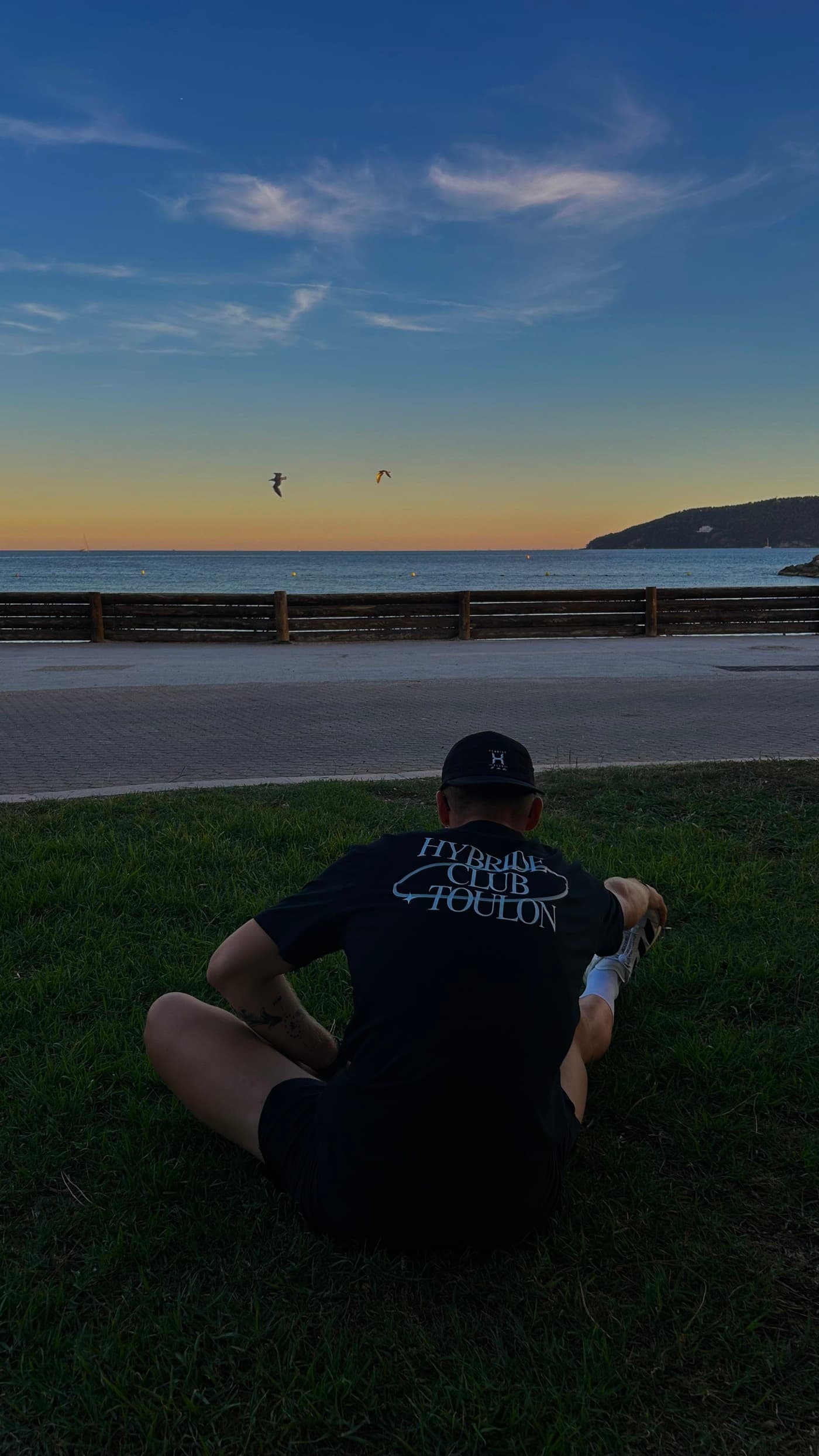 Un membre du club assis de dos dans l’herbe face à la mer, tee-shirt noir floqué Hybride Club Toulon