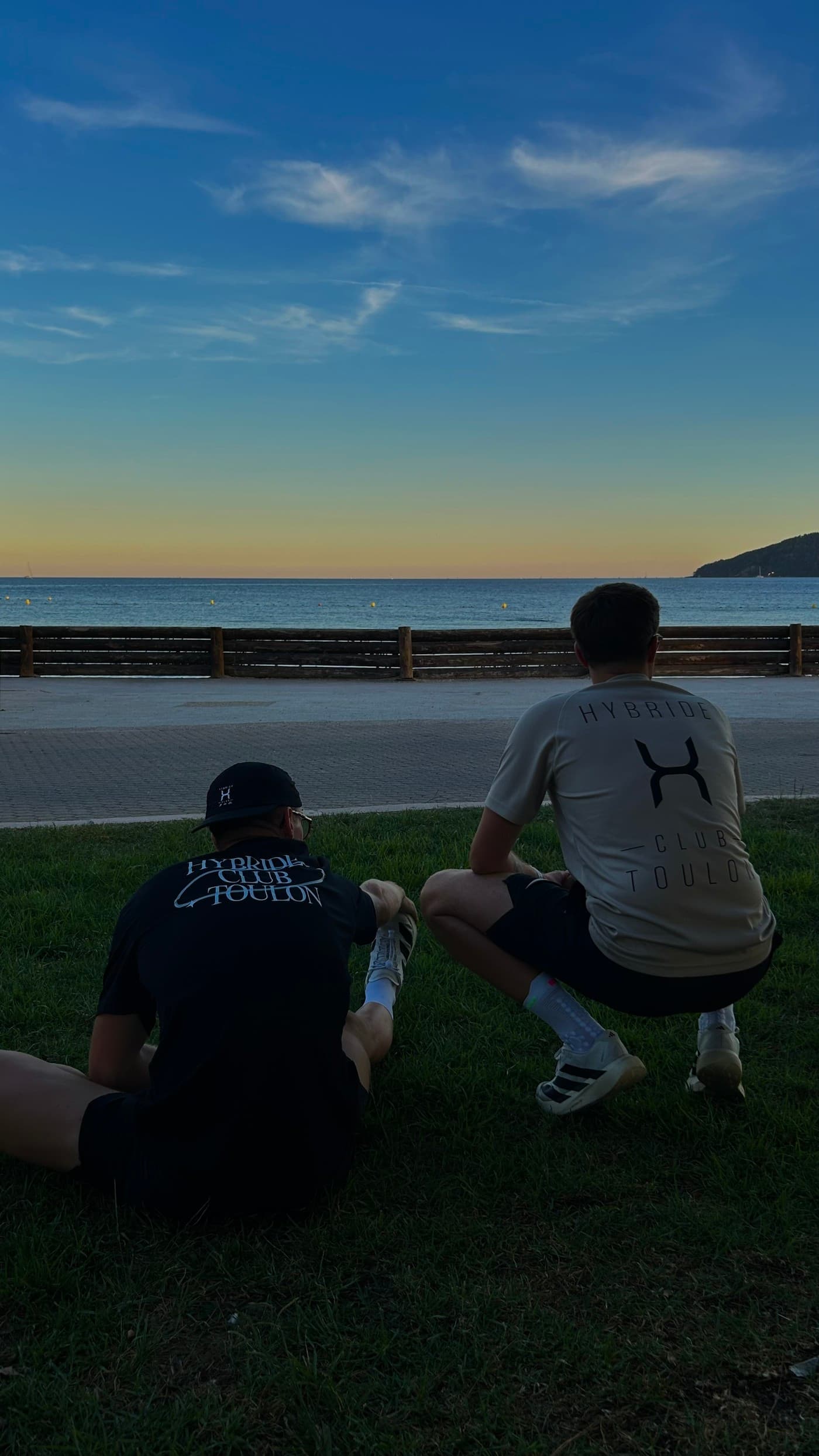 Deux membres du club de dos dans l’herbe face à la mer, tee-shirt noir floqué Hybride Club Toulon et tee-shirt beige au logo H