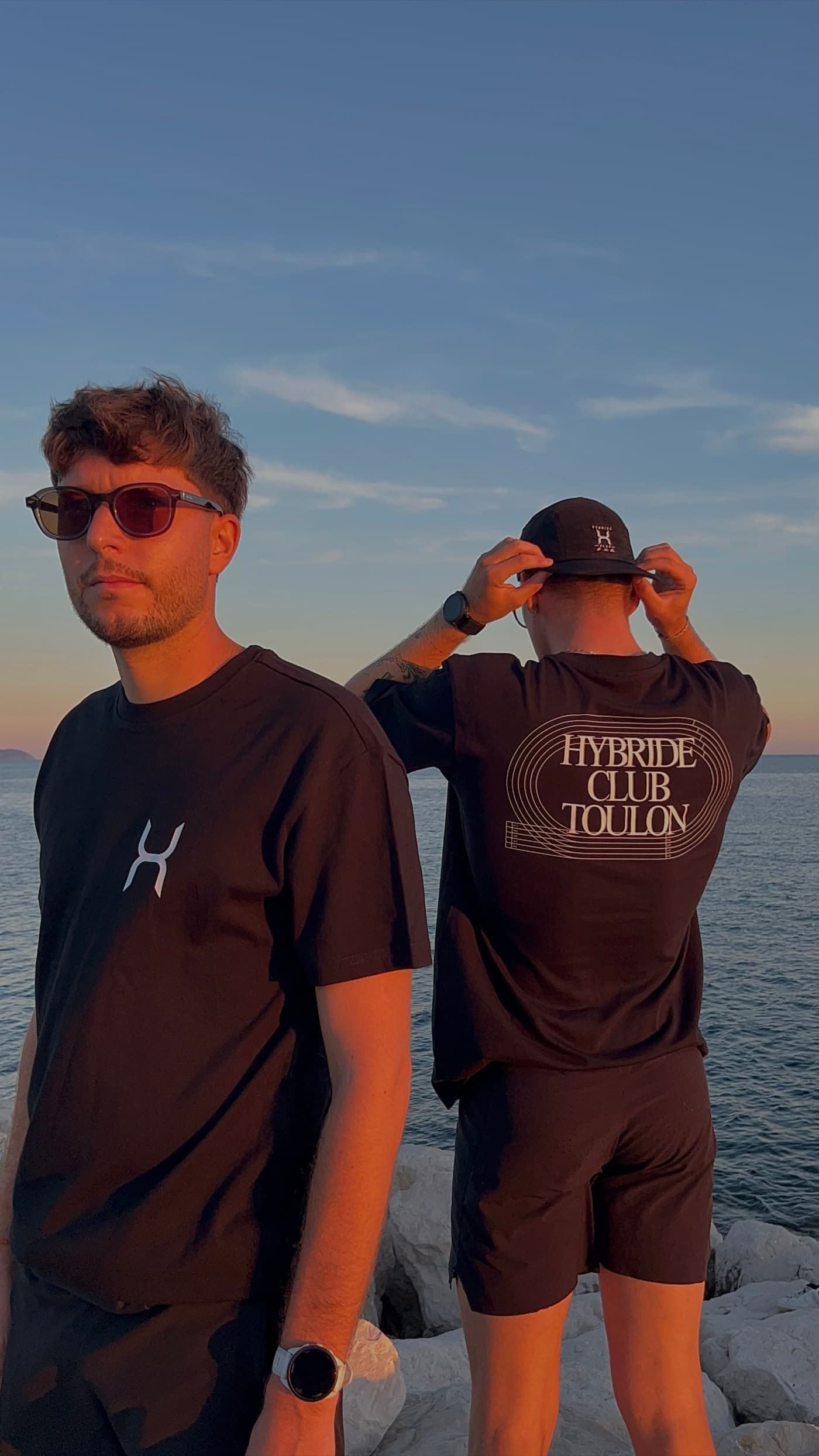 Deux membres du club au bord de mer : l’un de face, logo H sur le tee-shirt noir, l’autre de dos, inscription Hybride Club Toulon