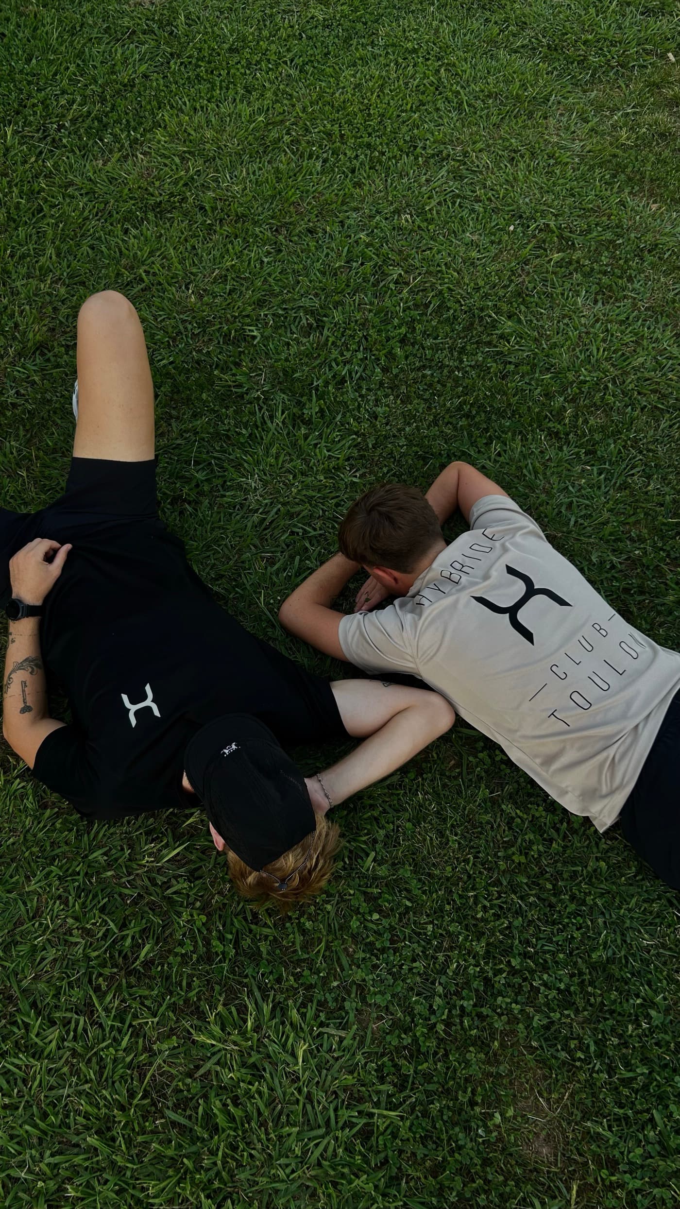Deux membres du club allongés dans l’herbe, l’un en tee-shirt noir au logo H, l’autre en tee-shirt crème floqué Hybride Club Toulon