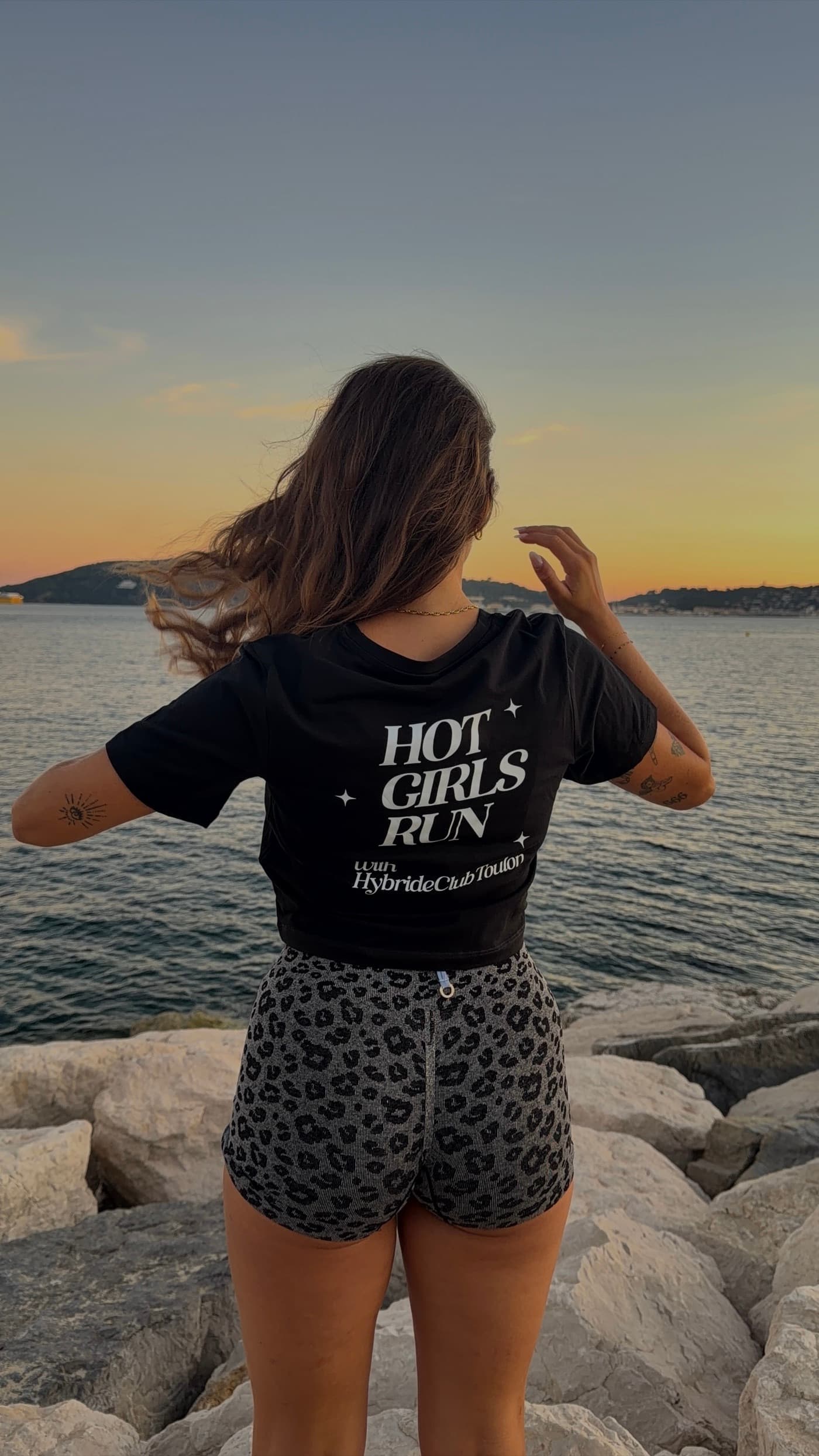 Une membre du club de dos au bord de mer, tee-shirt noir floqué Hot Girls Run
