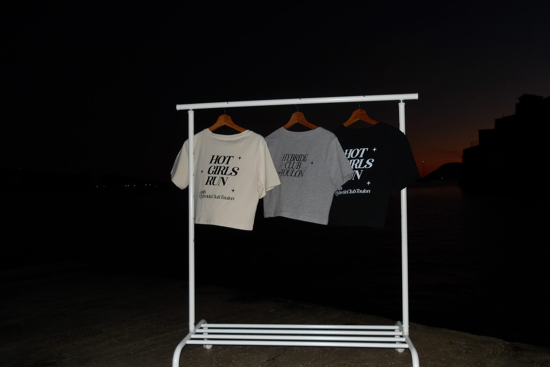 Trois tee-shirts du club sur un portant blanc, de nuit : crème et noir floqués Hot Girls Run, gris floqué Hybride Club Toulon