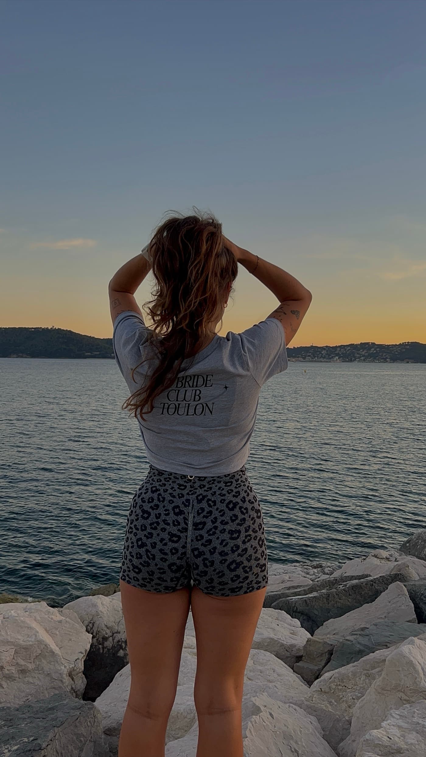 Une membre du club de dos face à la mer, en tee-shirt crème du club