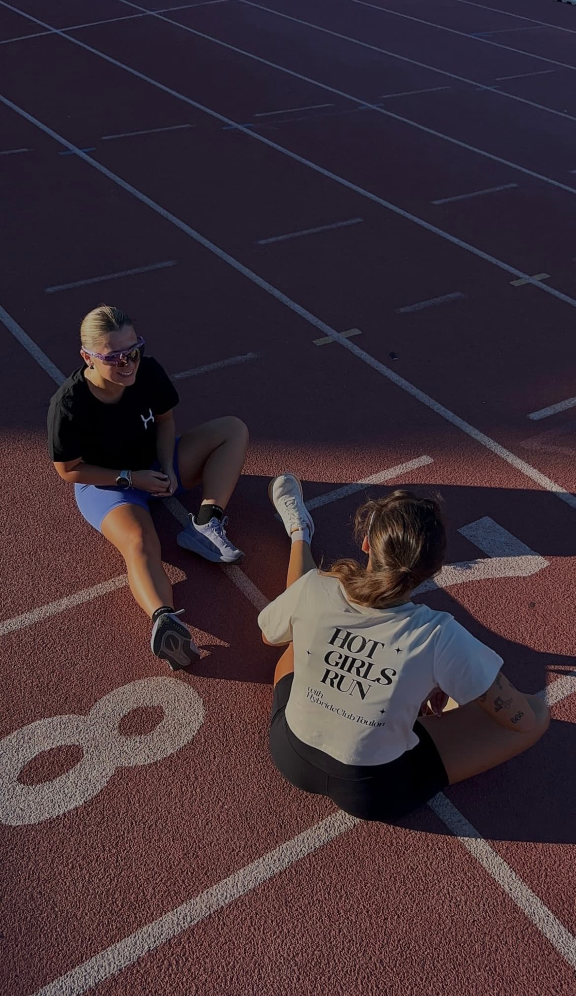 Deux membres du club assises sur la piste, tee-shirt noir et tee-shirt crème floqué Hot Girls Run