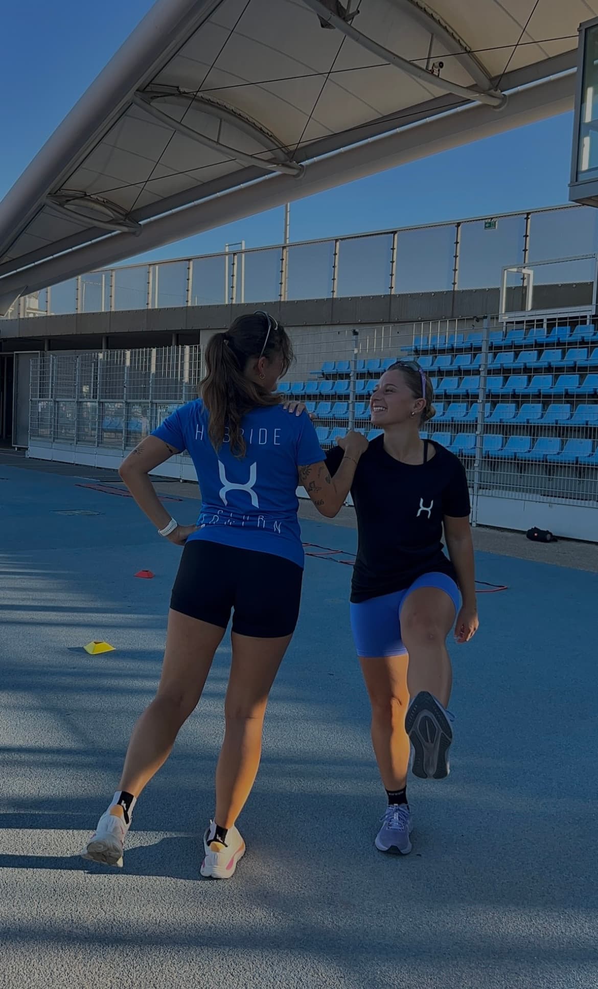 Deux membres du club debout sur la piste, tee-shirts bleu et noir au logo H, gradins en arrière-plan