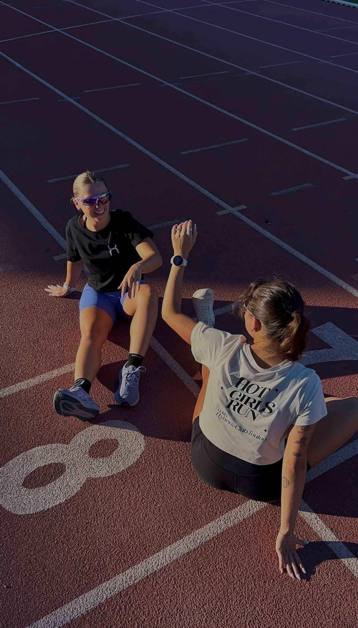 Deux membres du club en étirements sur la piste, tee-shirt noir et tee-shirt crème floqué Hot Girls Run