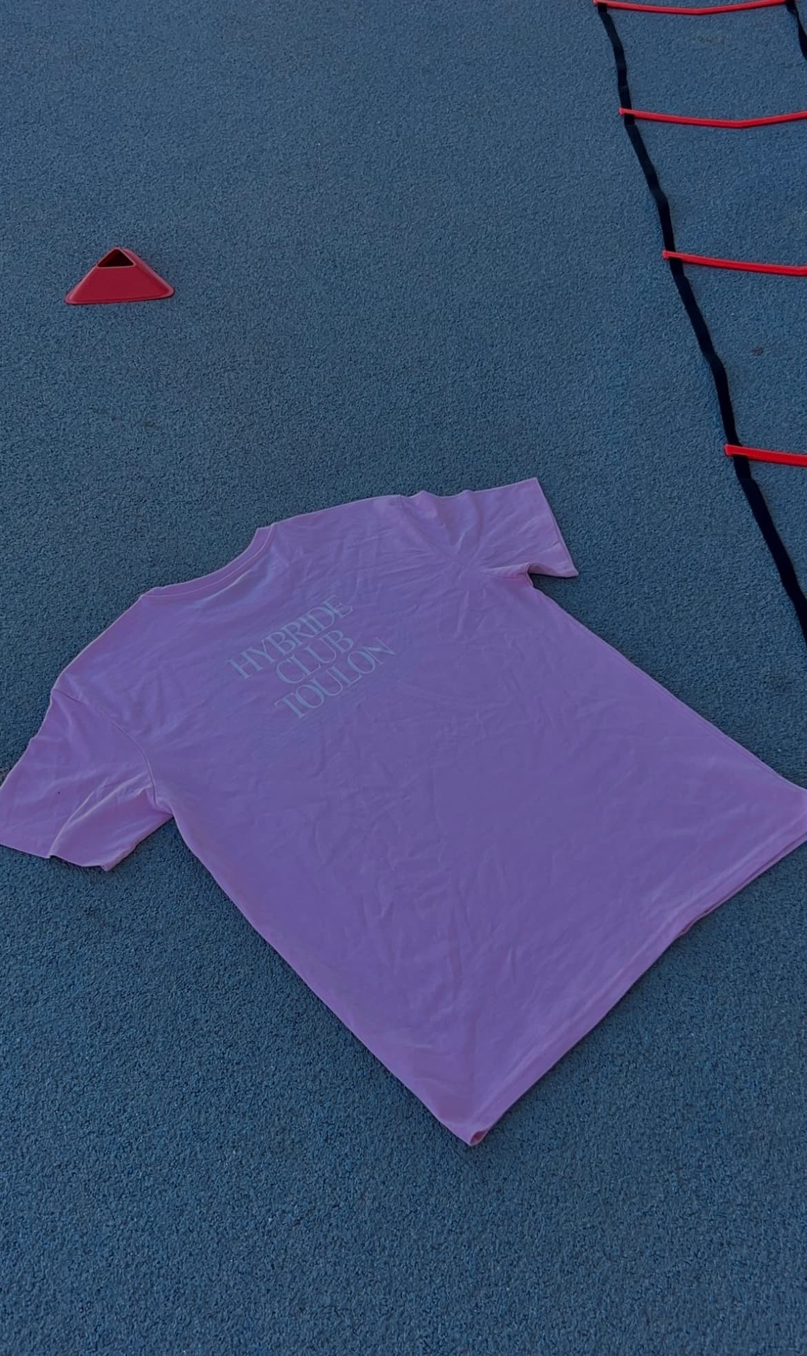 Un tee-shirt lavande du club posé à plat sur la piste d’athlétisme
